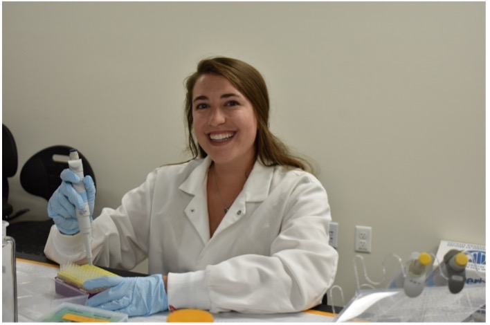 Tabor Whitney, a white woman, wearing a white lab coat and blue gloves. She is smiling. 