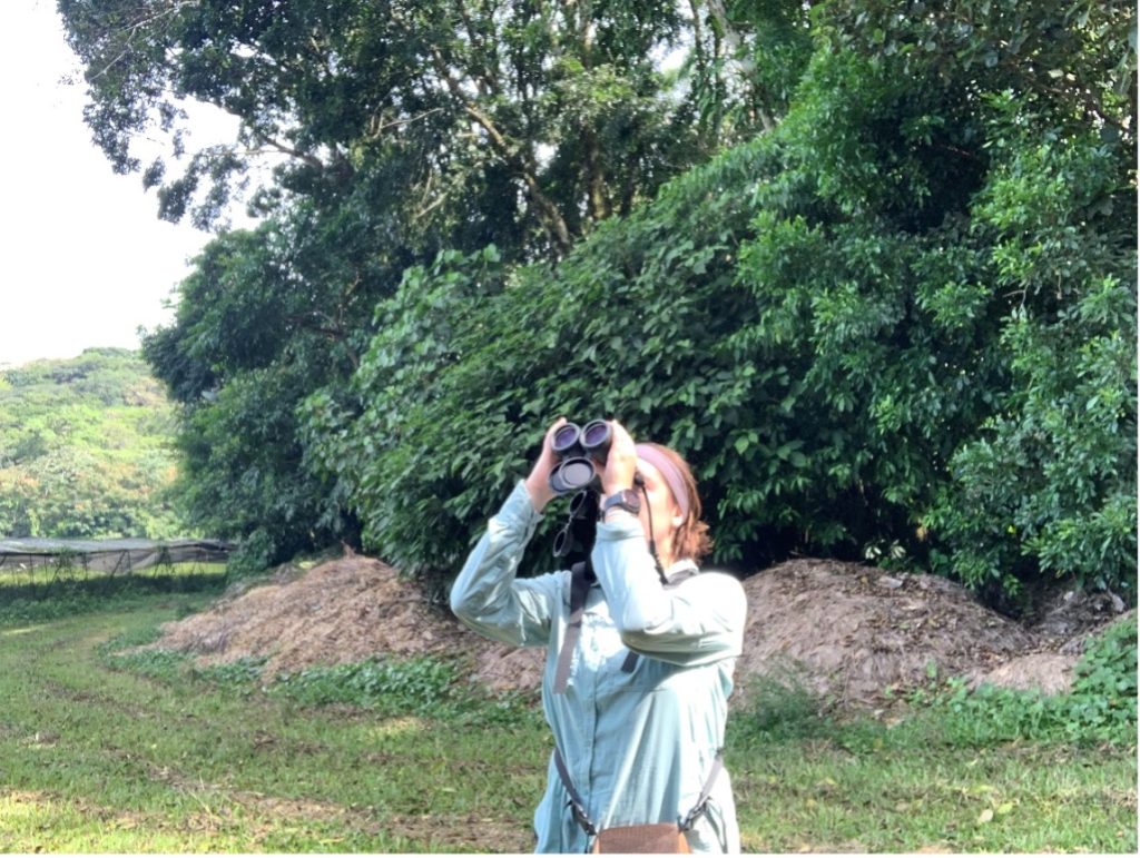 Tabor Whitney, a white woman, standing a field looking through binoculars